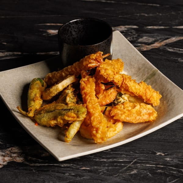 A plate of golden-brown tempura, including shrimp and vegetables, with a dark dipping cup.