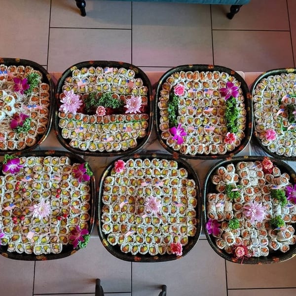 Six large platters of assorted sushi rolls, garnished with flowers and American flags.