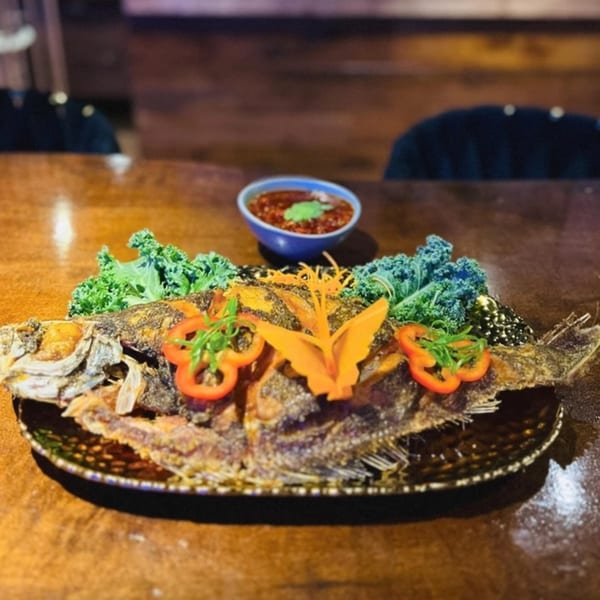 A whole fried fish, garnished with kale, bell peppers, and carrots, with a side of sauce.