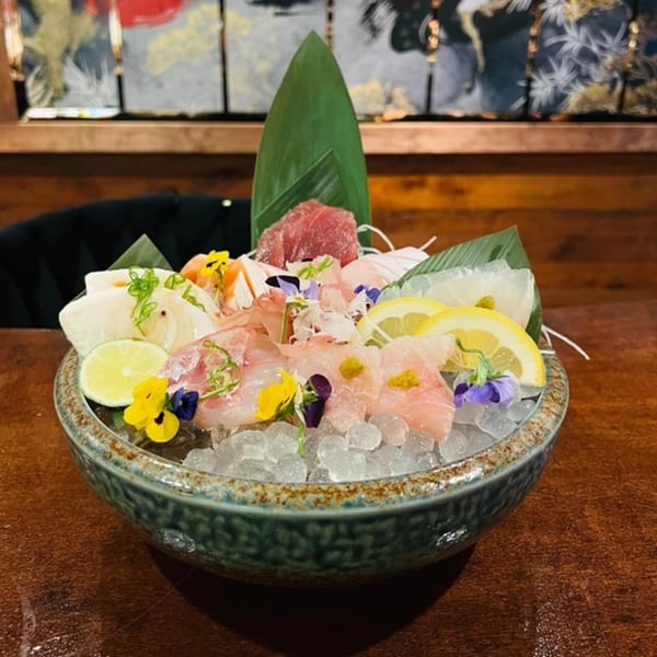 A colorful sashimi platter with various fish, lemon, and edible flowers, served on ice in a bowl.