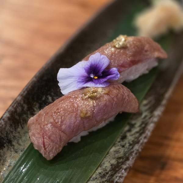 Two pieces of Akeno Sushi Bar and Thai wagyu nigiri, garnished with a purple flower and gold flakes.