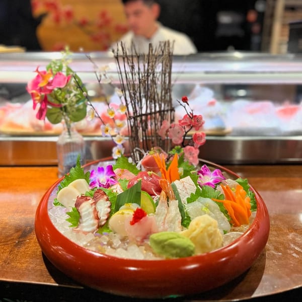 A large red bowl of assorted sashimi and seafood on ice, with a chef in the background.