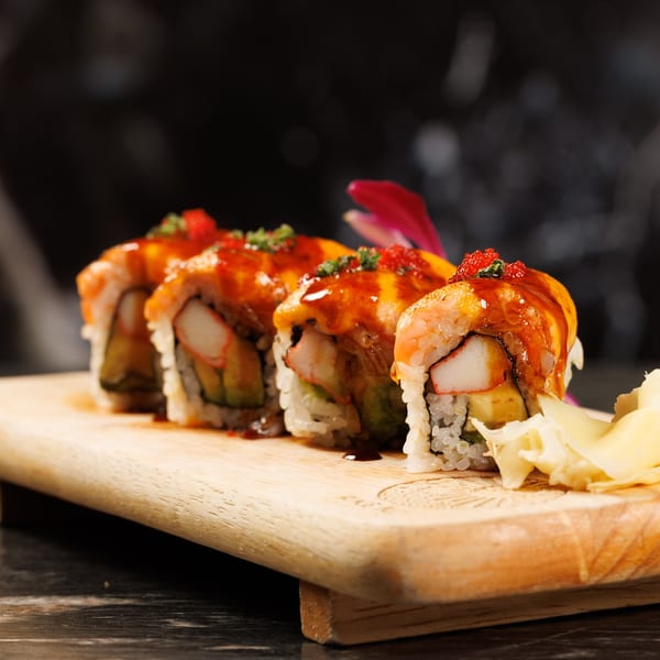 Four sushi rolls with crab, avocado, and egg, topped with sauce and roe on a wooden board.
