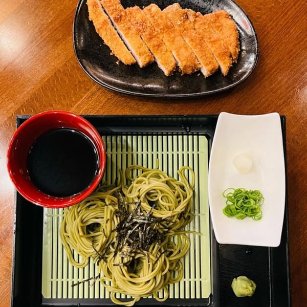 A plate of sliced tonkatsu and a tray of green soba noodles with dipping sauce and garnishes.