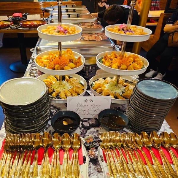 A buffet table with tiered platters of fried appetizers, sauces, stacks of plates, and golden cutlery.
