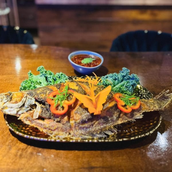 Whole fried fish garnished with kale, bell peppers, and carrot, with a side of sauce.