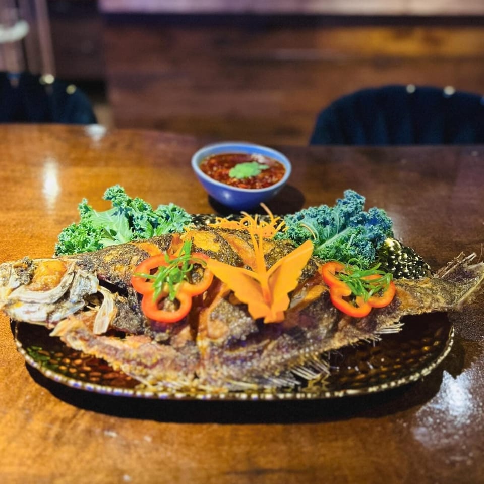 Whole fried fish garnished with kale, bell peppers, and carrot, with a side of sauce.